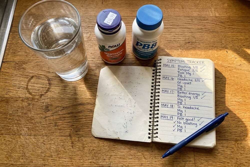 Overhead kitchen counter with a glass of water, a magnesium bottle, a probiotic bottle, and a small notebook tracking bloating symptoms across days in warm afternoon light