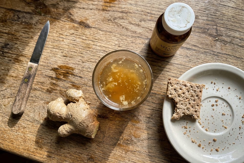 Overhead view of a kitchen counter at 7am with a half-eaten sleeve of saltine crackers, ginger tea steeping in a mug, and a vitamin B6 bottle on a wood surface