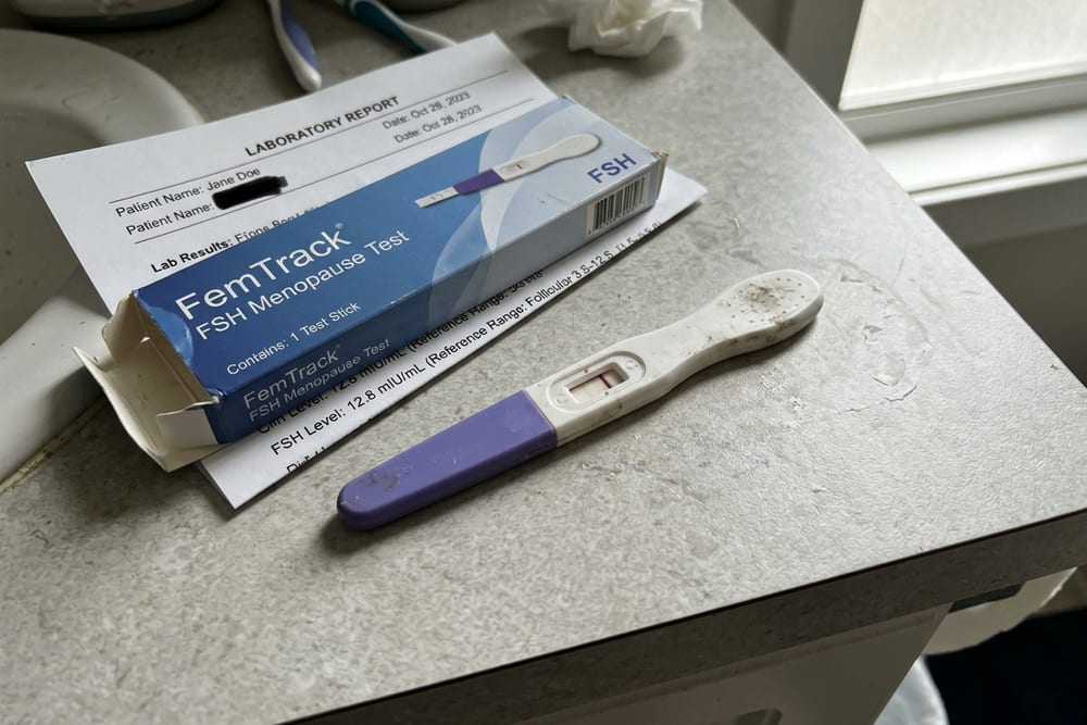 Overhead close-up of an at-home FSH urine test stick on a bathroom counter next to its box and a printed lab results sheet from a doctor's office